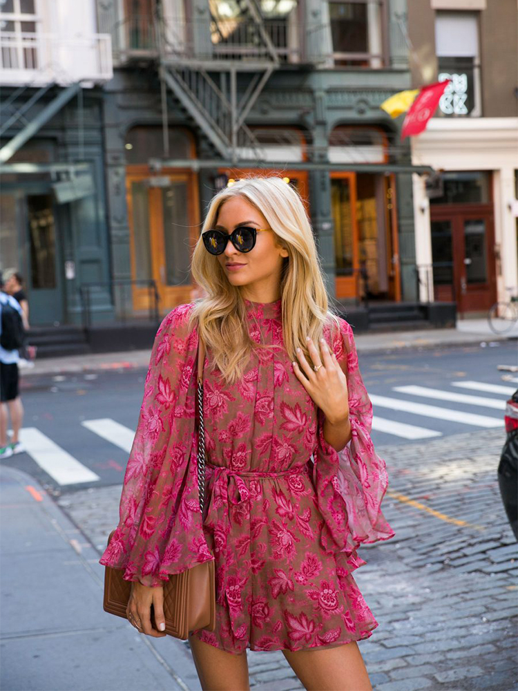 Femme blonde marchant dans la rue portant une combinaison rose florale avec un sac sur son épaule droite et une paire de lunette de soleil. Derrière elle se trouve un passage piéton.